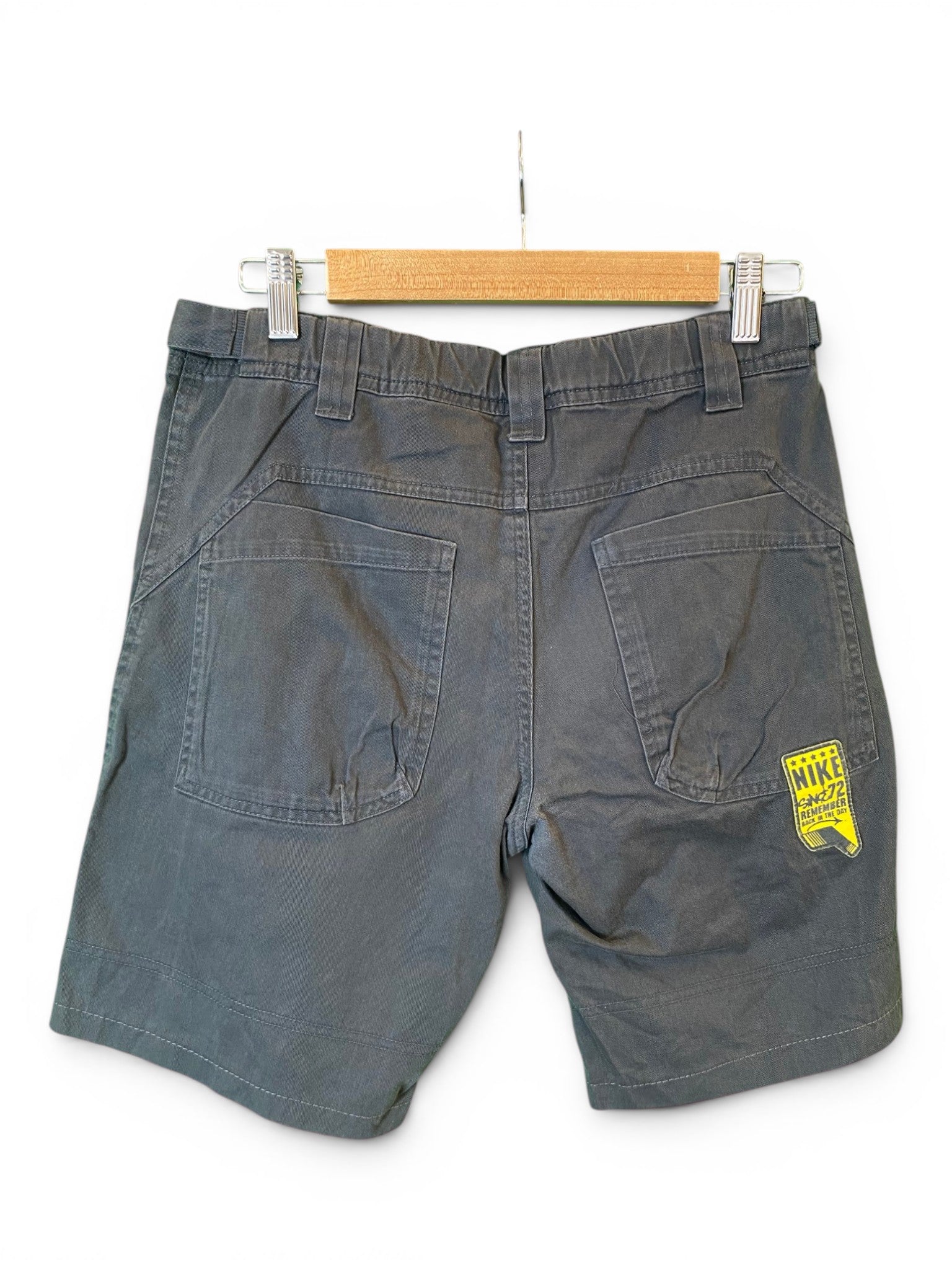 A pair of dark gray denim shorts hanging on a wooden hanger against a white background, showing the back pockets and a yellow Nike Swoosh logo patch on the right pocket.