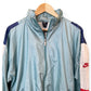 A light blue Nike windbreaker jacket hanging on a wooden hanger against a white background, featuring navy blue shoulder panels, white lower sleeves, and a red Nike logo.