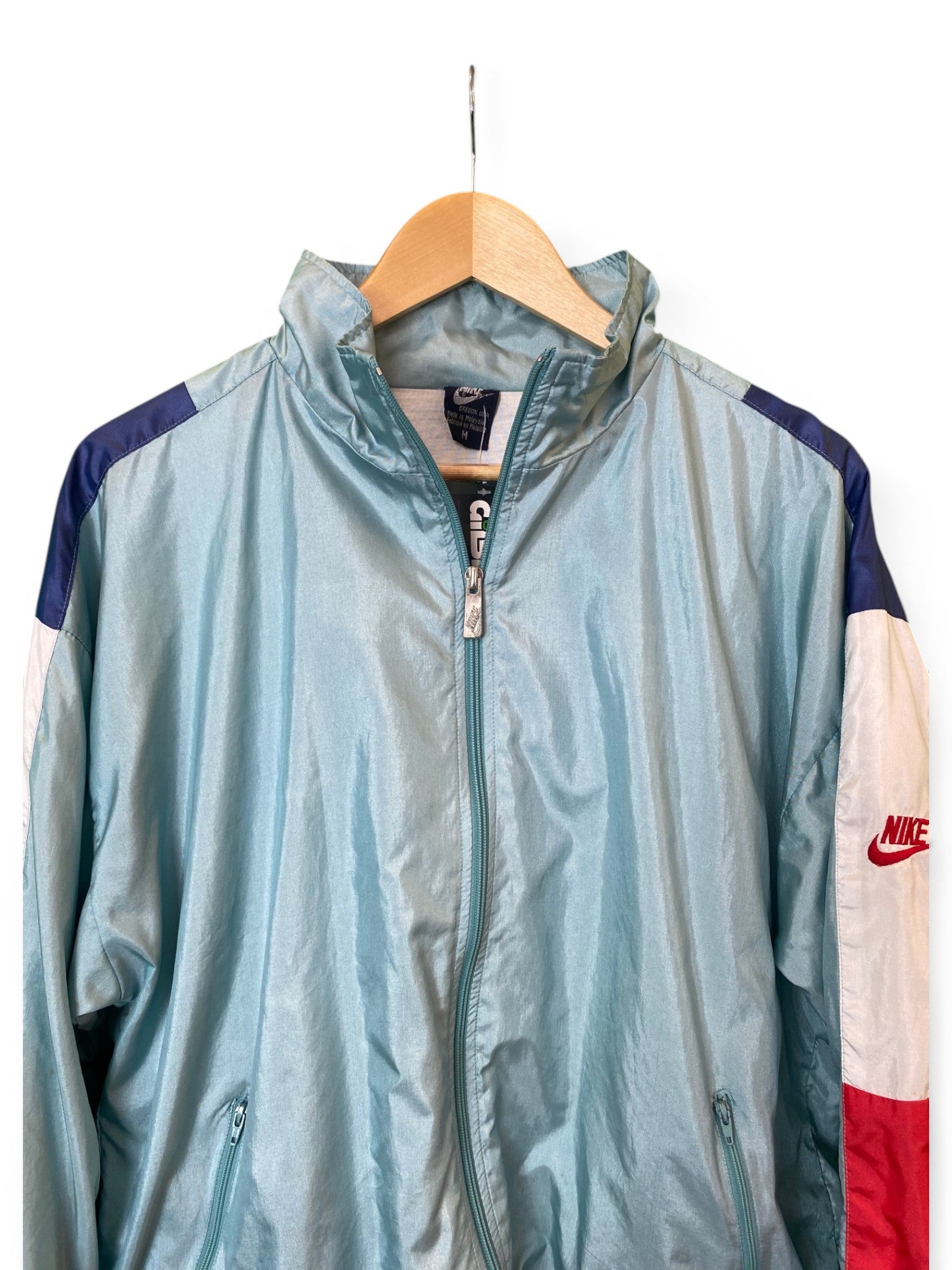 A light blue Nike windbreaker jacket hanging on a wooden hanger against a white background, featuring navy blue shoulder panels, white lower sleeves, and a red Nike logo.